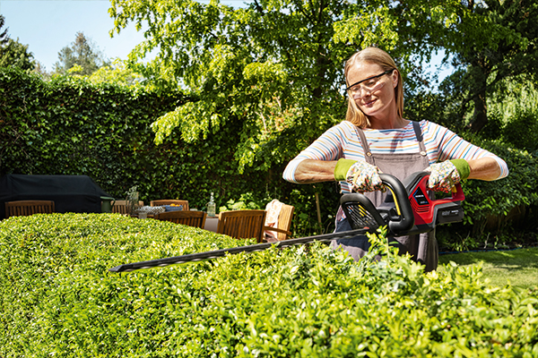 Eine Frau mit Schutzbrille schneidet die Hecke in ihrem Garten mit einem Honda Akku-Rasentrimmer.